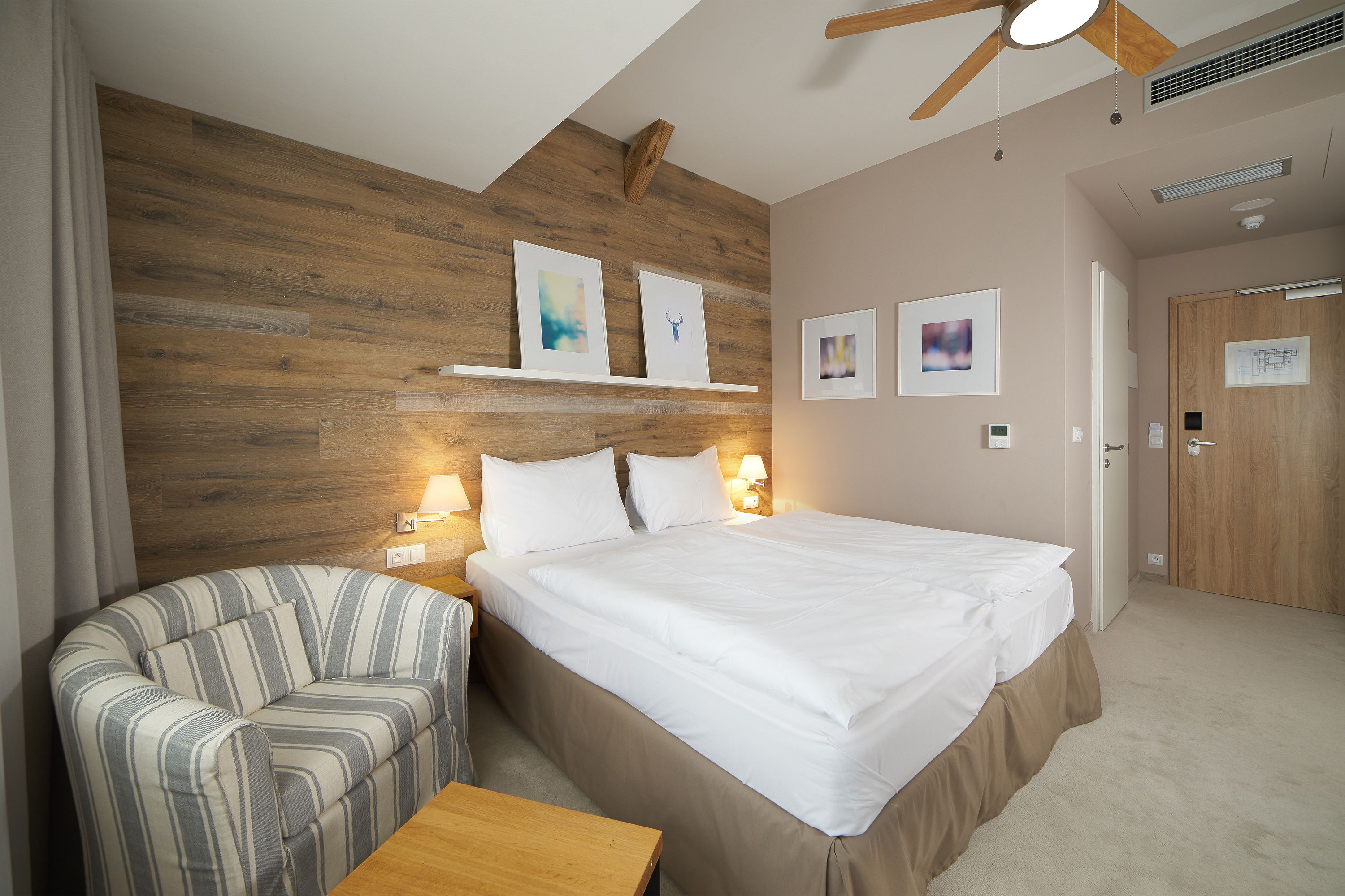 Una habitación de hotel con una cama grande, un sillón a rayas, una mesita de madera y un ventilador de techo.