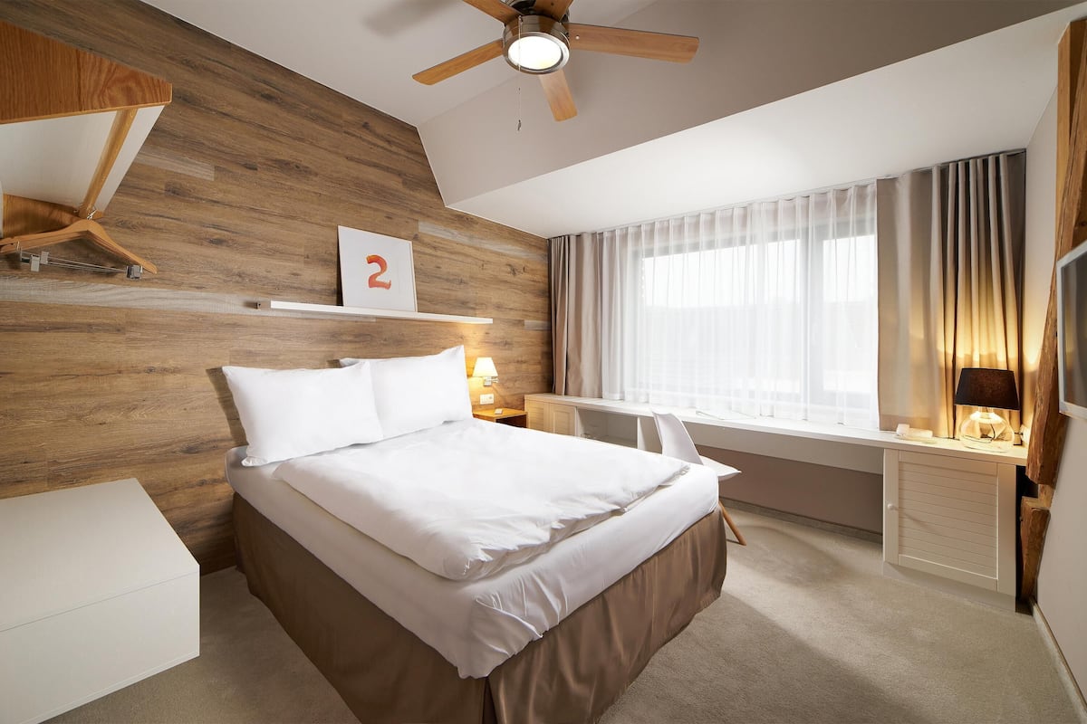Een moderne hotelkamer met een houten accentmuur, een groot bed, een bureau met stoel, een plafondventilator en een groot raam met gordijnen.
