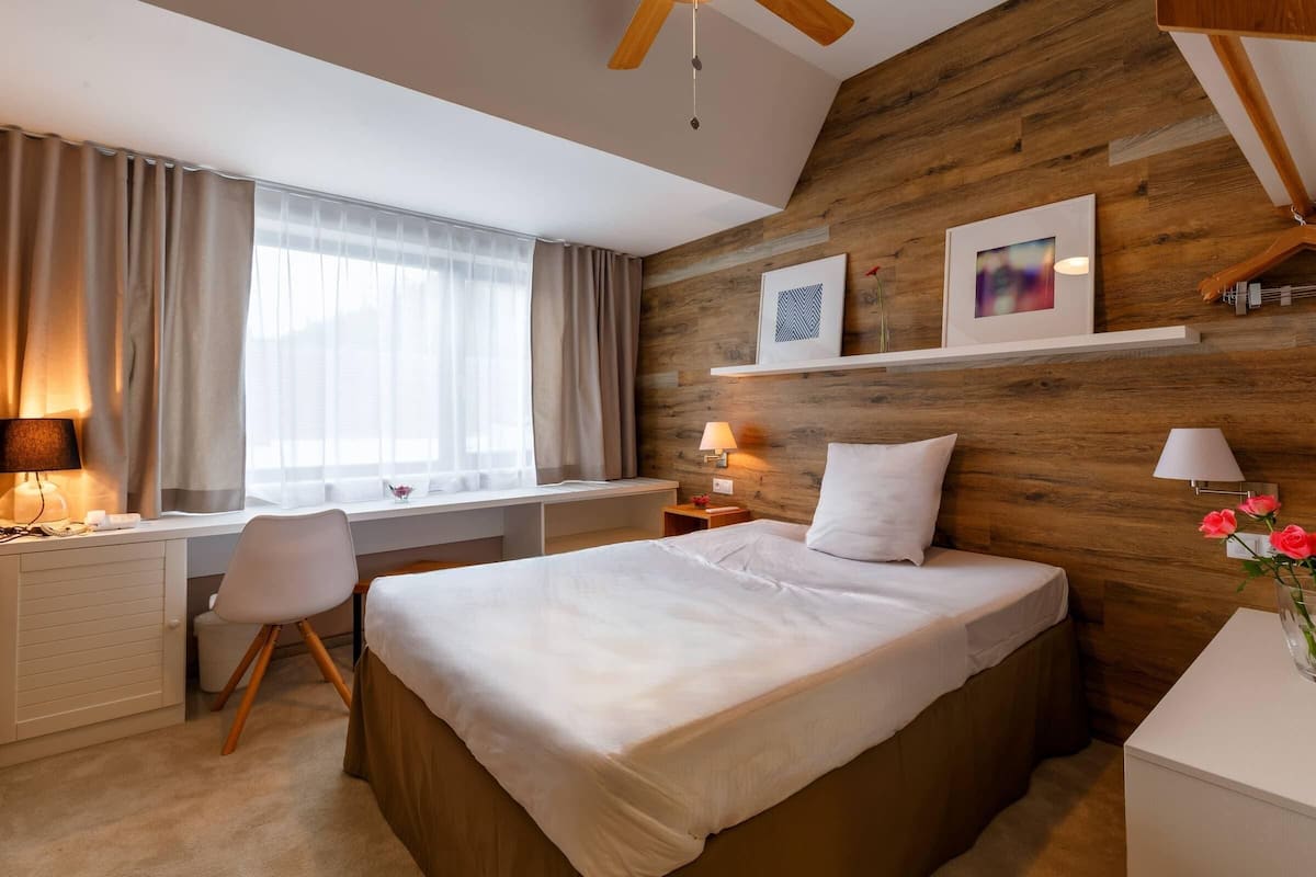 Een slaapkamer met een wand van hout, een bureau met stoel, een bed met wit beddengoed en een nachtkastje met lamp.