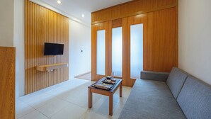 A modern living room with a wooden panel wall, a flat-screen TV mounted on the wall, a grey sofa, and a wooden coffee table. - Bali True Living Apartment (Denpasar)