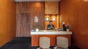 A reception area with a wooden panel wall, a receptionist, and two wicker stools. - Bali True Living Apartment (Denpasar)