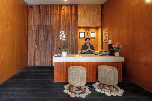A reception area with a wooden panel wall, a receptionist, and two wicker stools. - Bali True Living Apartment (Denpasar)