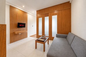A modern living room with a grey sofa, wooden panel walls, a flat-screen TV, and a small wooden table with a tray. - Bali True Living Apartment (Denpasar)