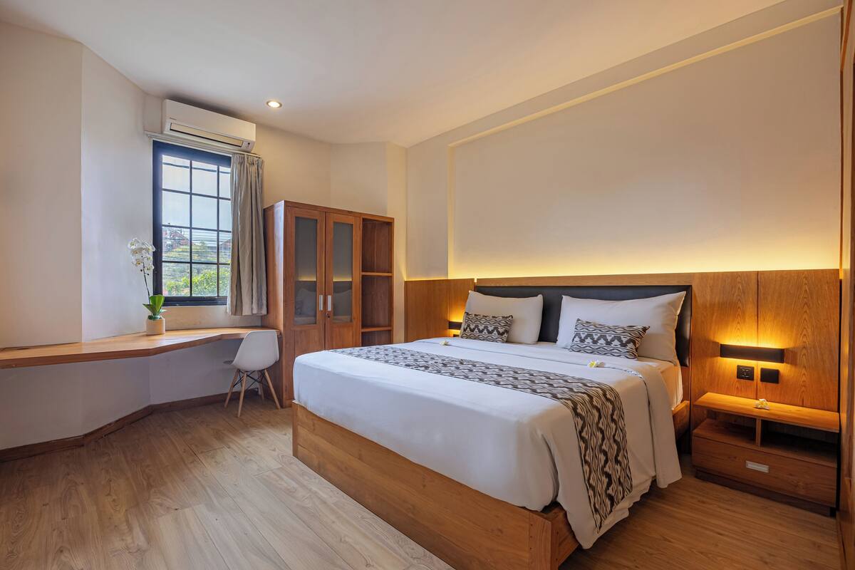 A bedroom with a large bed, a desk, a wardrobe, and a bedside table with a lamp.