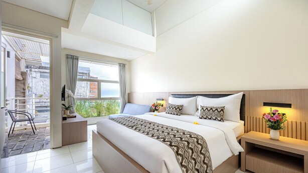 A modern hotel room with a large bed, a bedside table with a lamp, a small desk, and a view of the outside balcony. - Bali True Living Apartment (Denpasar)