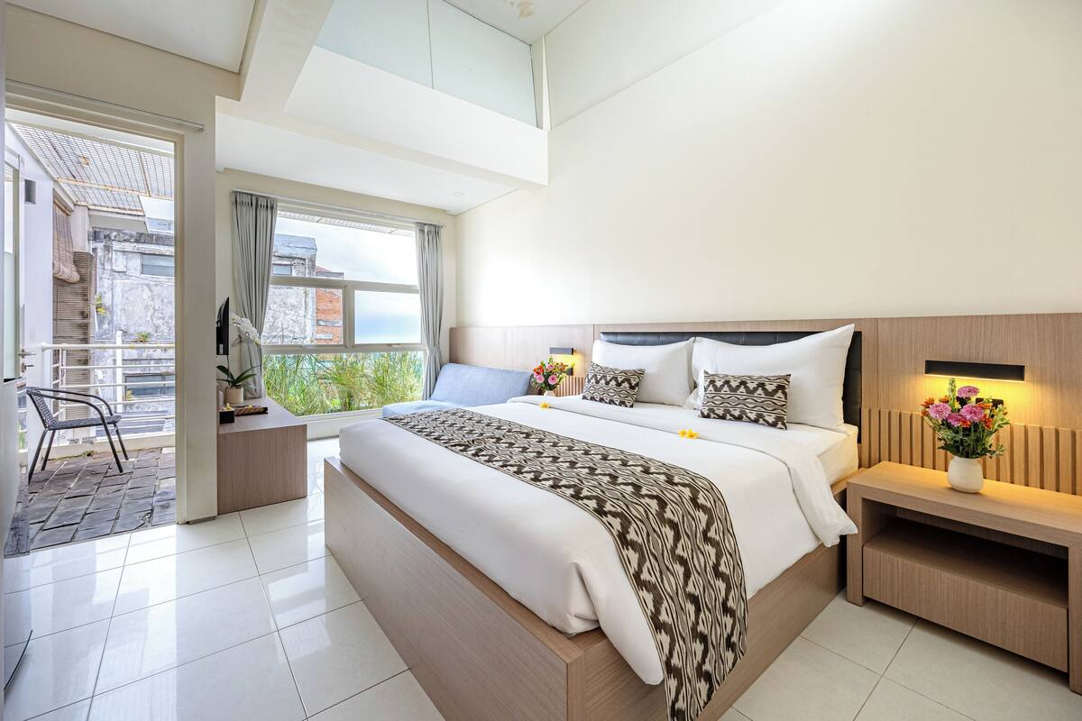 A modern hotel room with a large bed, a bedside table with a lamp, a small desk, and a view of the outside balcony.