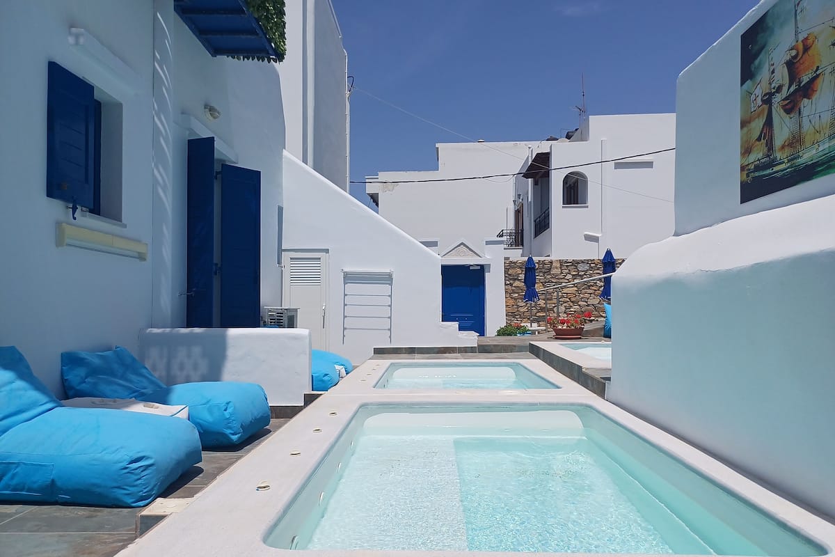 Un área de piscina con tumbonas azules y una piscina cristalina, rodeada de edificios blancos con detalles azules.