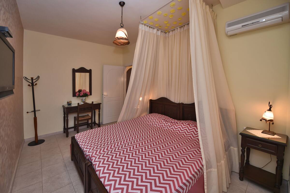 Un dormitorio con una cama, una mesita de noche con lámpara, un escritorio con silla, un perchero y un aire acondicionado instalado en la pared.