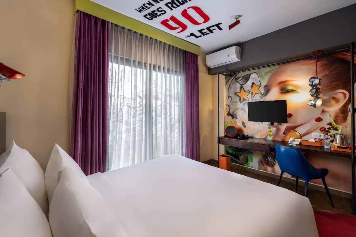 A hotel room with a bed, a desk with a computer, a chair, and a wall with a mural.