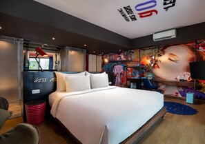 A modern hotel room with a large bed, a red trash bin, and a wall with colorful artwork.