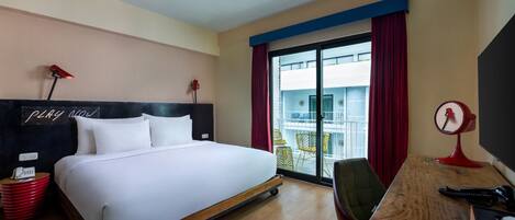 A hotel room with a bed, a desk with a chair, a red lamp, and a red clock.