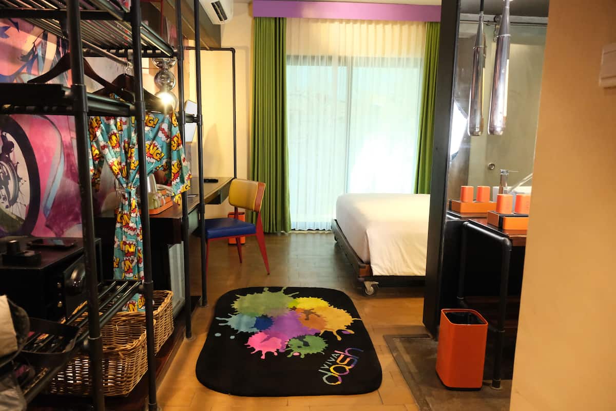 A hotel room with a bed, a desk, a chair, a colorful rug, and a window with curtains.