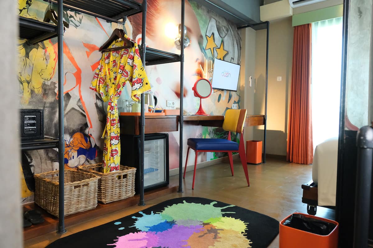 A room with a colorful abstract mural, a desk with a computer, a chair, and a small table with a lamp.