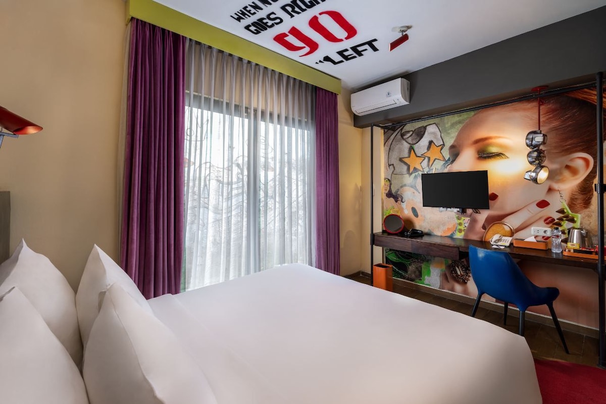 A hotel room with a bed, a desk with a computer, a chair, and a wall with a mural.