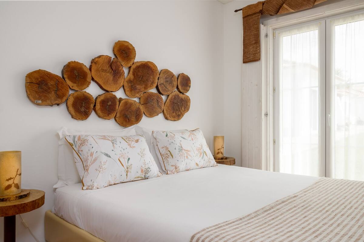 Un dormitorio con una cama grande, ropa de cama blanca, dos almohadas decorativas, una obra de arte de madera en la pared, una lámpara de noche y una ventana con cortina.