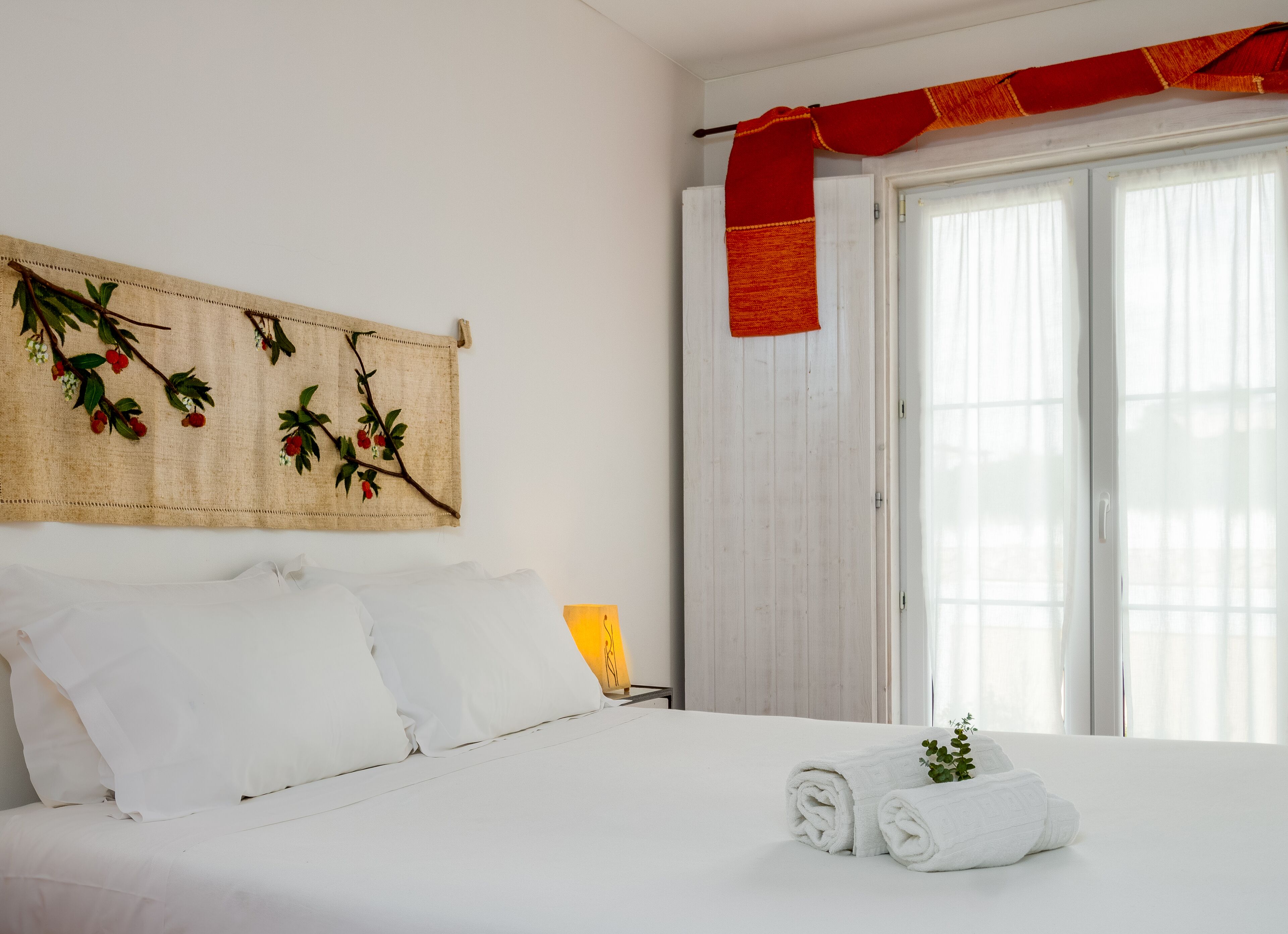 Una cama impecablemente tendida con sábanas blancas, un paño decorativo rojo en la ventana y una rama con bayas rojas montada en la pared.