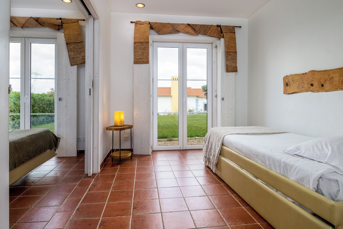 A bedroom with a bed, a bedside table with a lamp, a window with curtains, and a view of the outside.