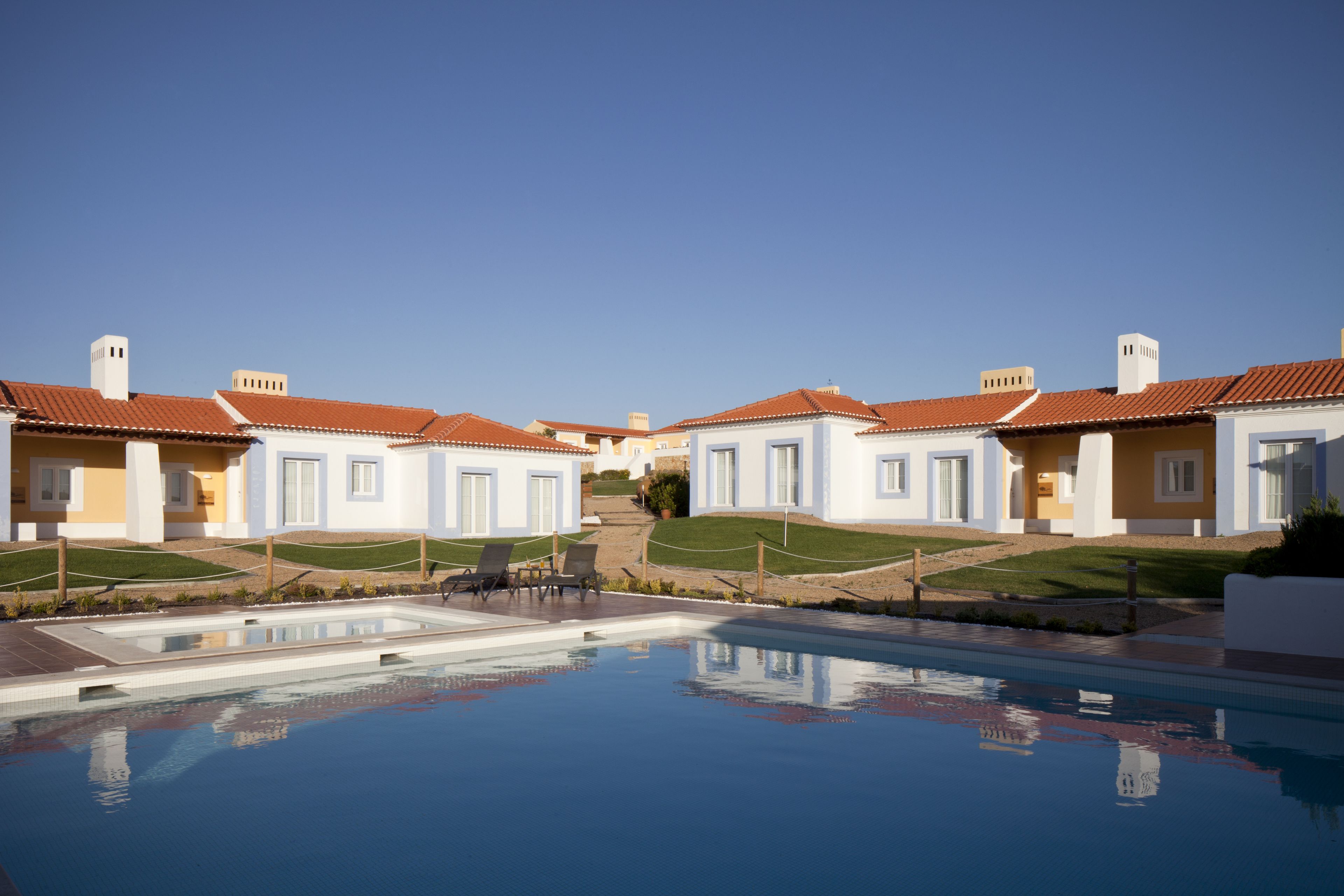 Un área de piscina con una piscina azul cristalina, rodeada de edificios blancos con techos rojos y un cielo despejado.
