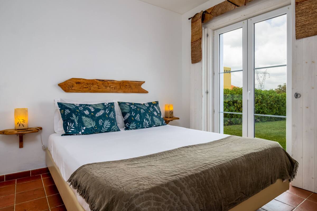 Un dormitorio con cama, dos lámparas de noche, una obra de arte de madera en la pared y vistas a zonas verdes a través de una puerta corrediza de vidrio.