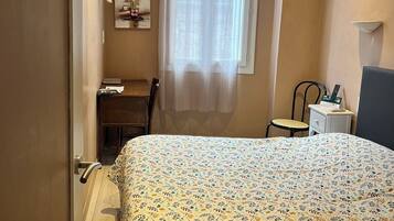 A bedroom with a bed, a desk, a chair, a nightstand, and a window with curtains.