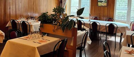 Zona pranzo caratterizzata da pannelli in legno, tavoli apparecchiati e ampie finestre con tende.