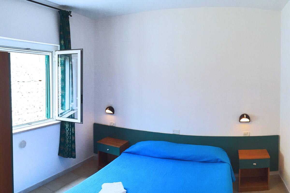 A bedroom with a single bed, two bedside tables, a window with curtains, and wall-mounted lights.