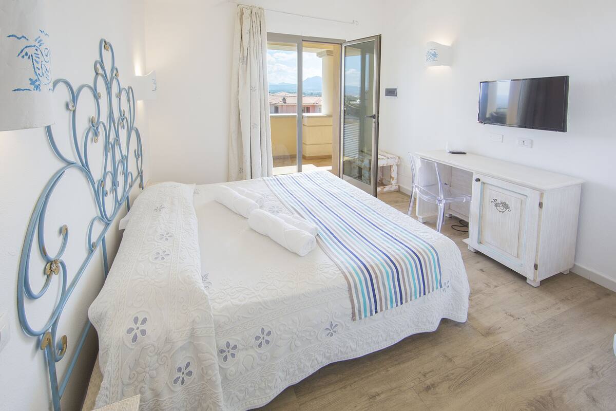 A bedroom with a bed, a desk, a TV, and a balcony with a view.