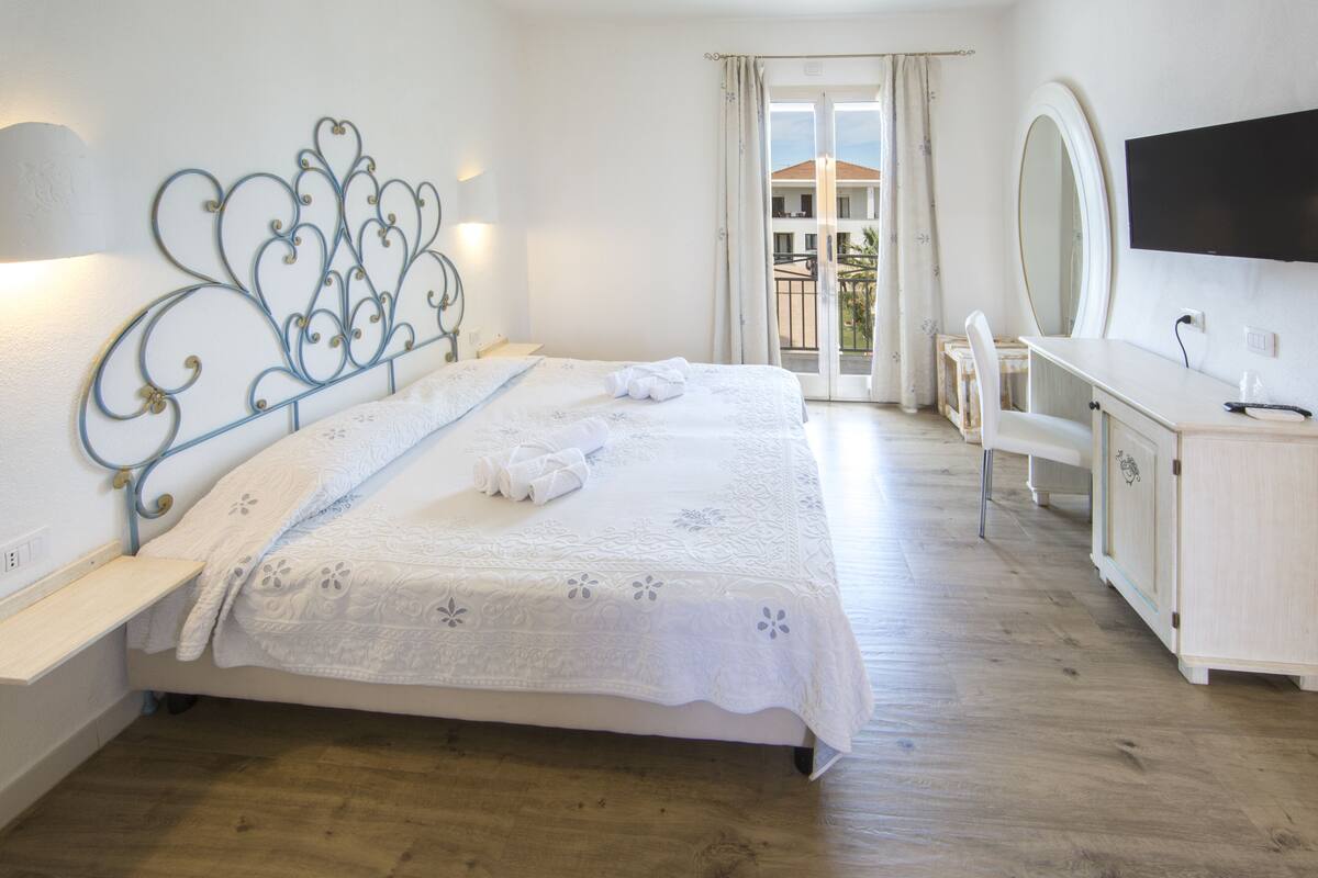 A bedroom with a bed, bedside table, dresser, TV, and a balcony with a view.