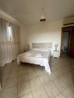 A bedroom with a bed, two bedside tables, a hanging light fixture, a patterned headboard, and a door leading to another room.