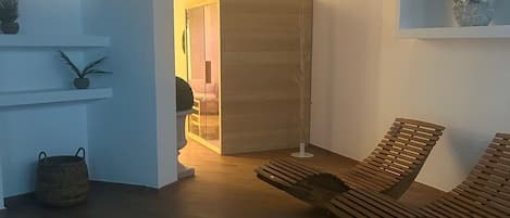 Un área de piscina interior moderna con una silla de descanso de madera y una pequeña mesa, rodeada de paredes blancas y un techo con follaje artificial.