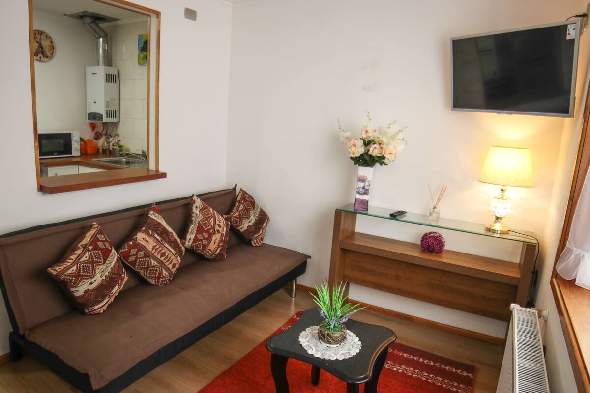 Una sala de estar con un sofá marrón, una mesa de centro de vidrio, un florero con flores, una lámpara y un televisor de pantalla plana montado en la pared.