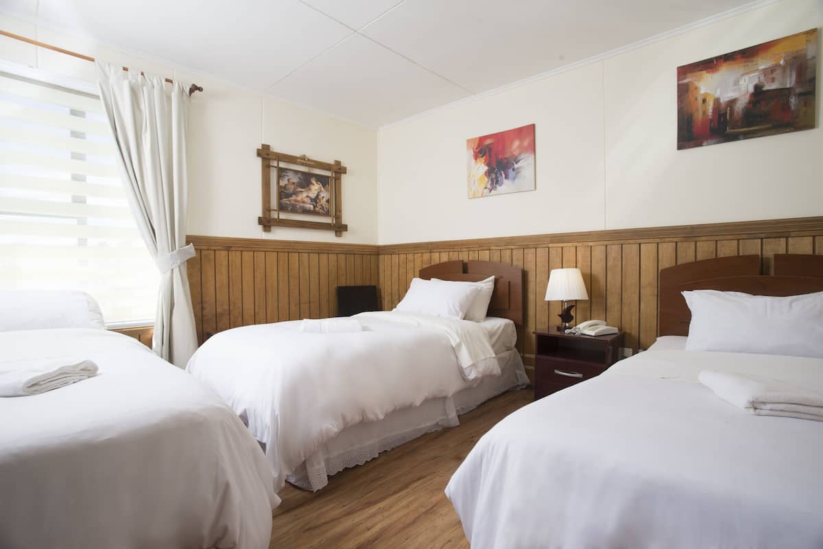 Habitación de hotel con tres camas, paneles de madera, un cuadro y una ventana con persianas.