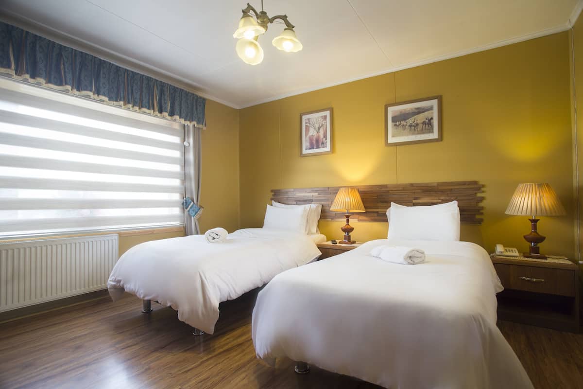 Habitación de hotel con dos camas, cabecero de madera, mesita de noche con lámpara y ventana con persianas.