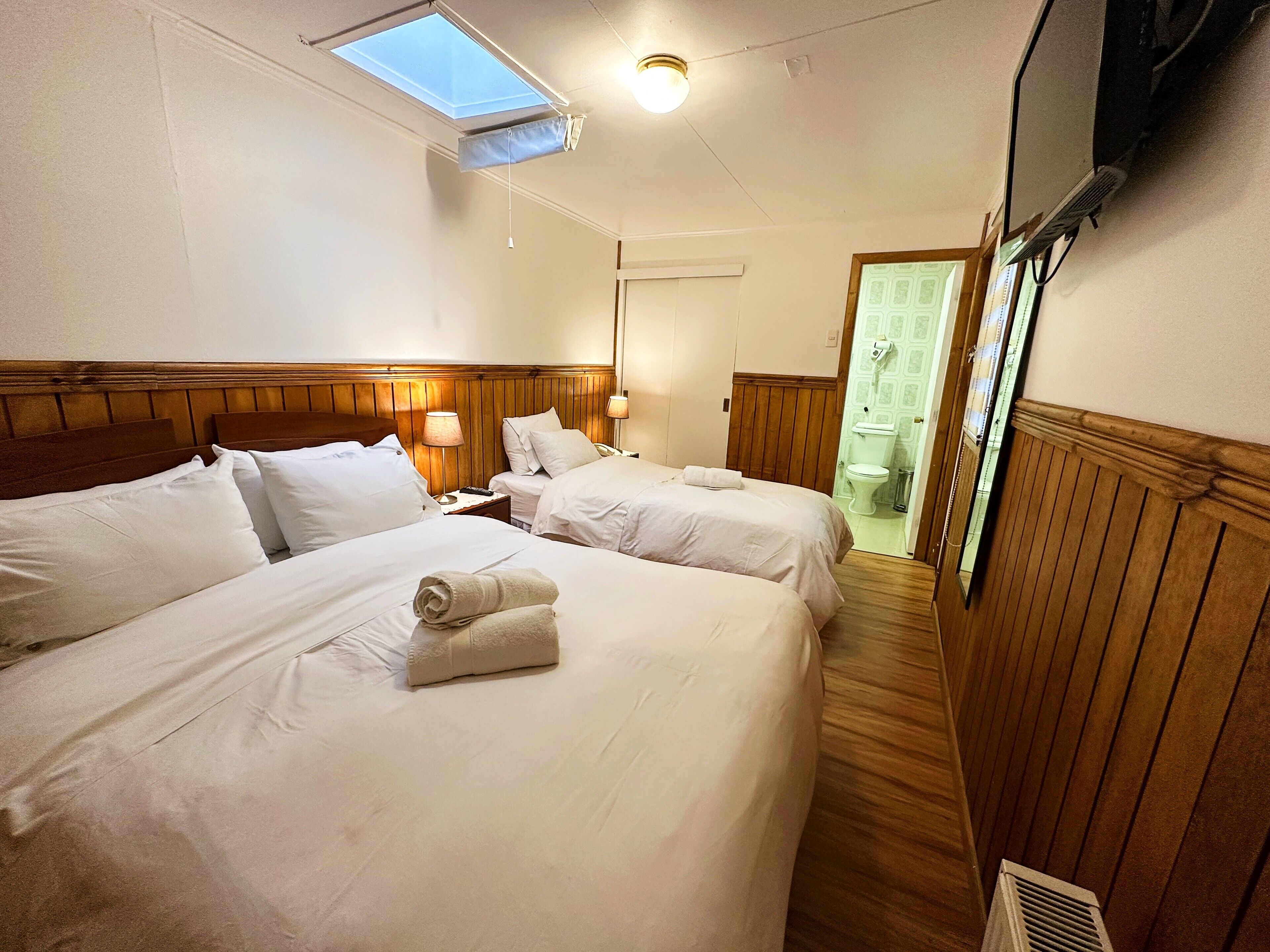 Habitación de hotel con dos camas, un tragaluz, un televisor y un baño visible a través de una puerta abierta.