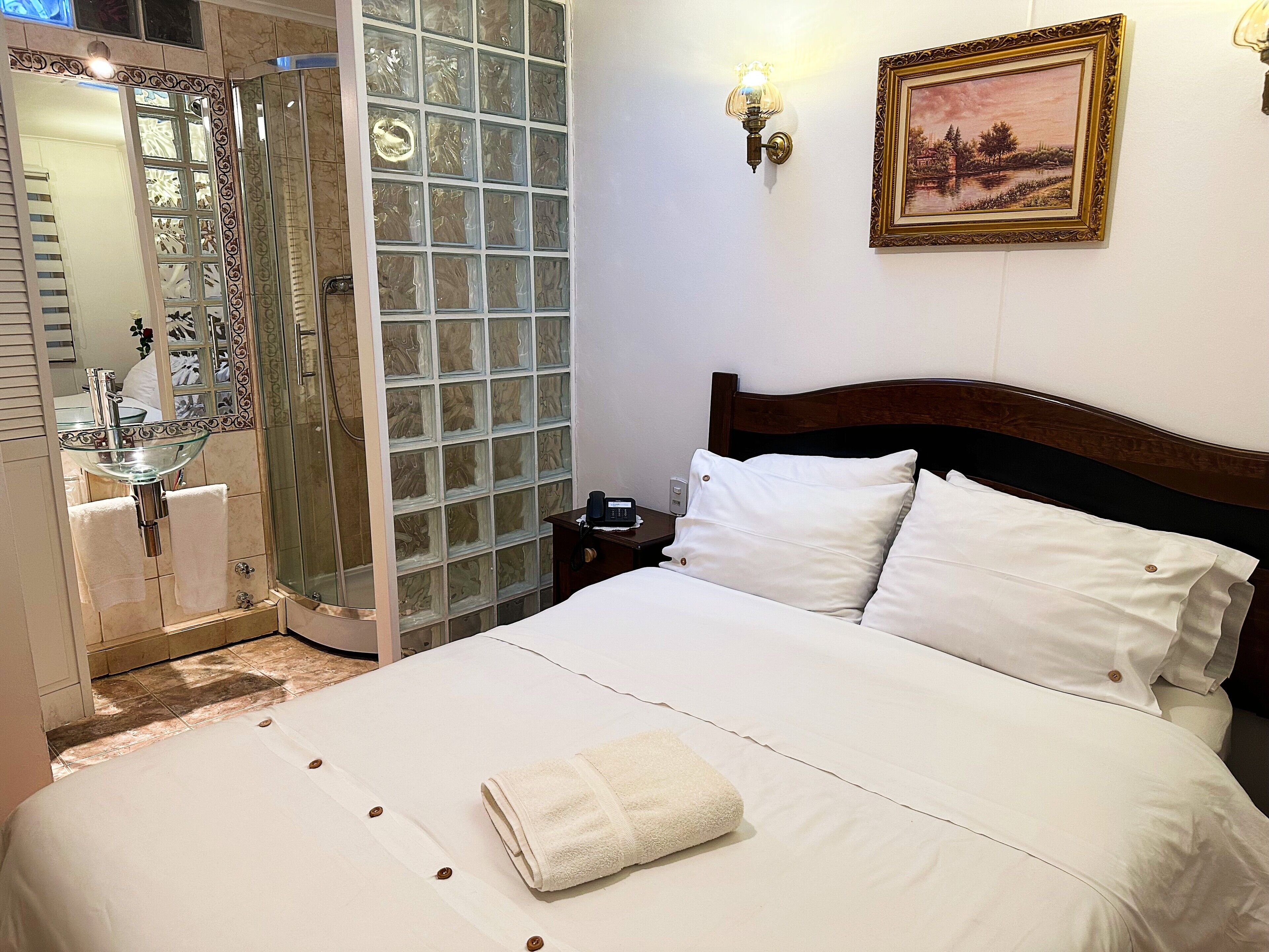 A hotel room with a bed, a nightstand, a painting, a glass shower enclosure, and a bathroom with a glass sink.