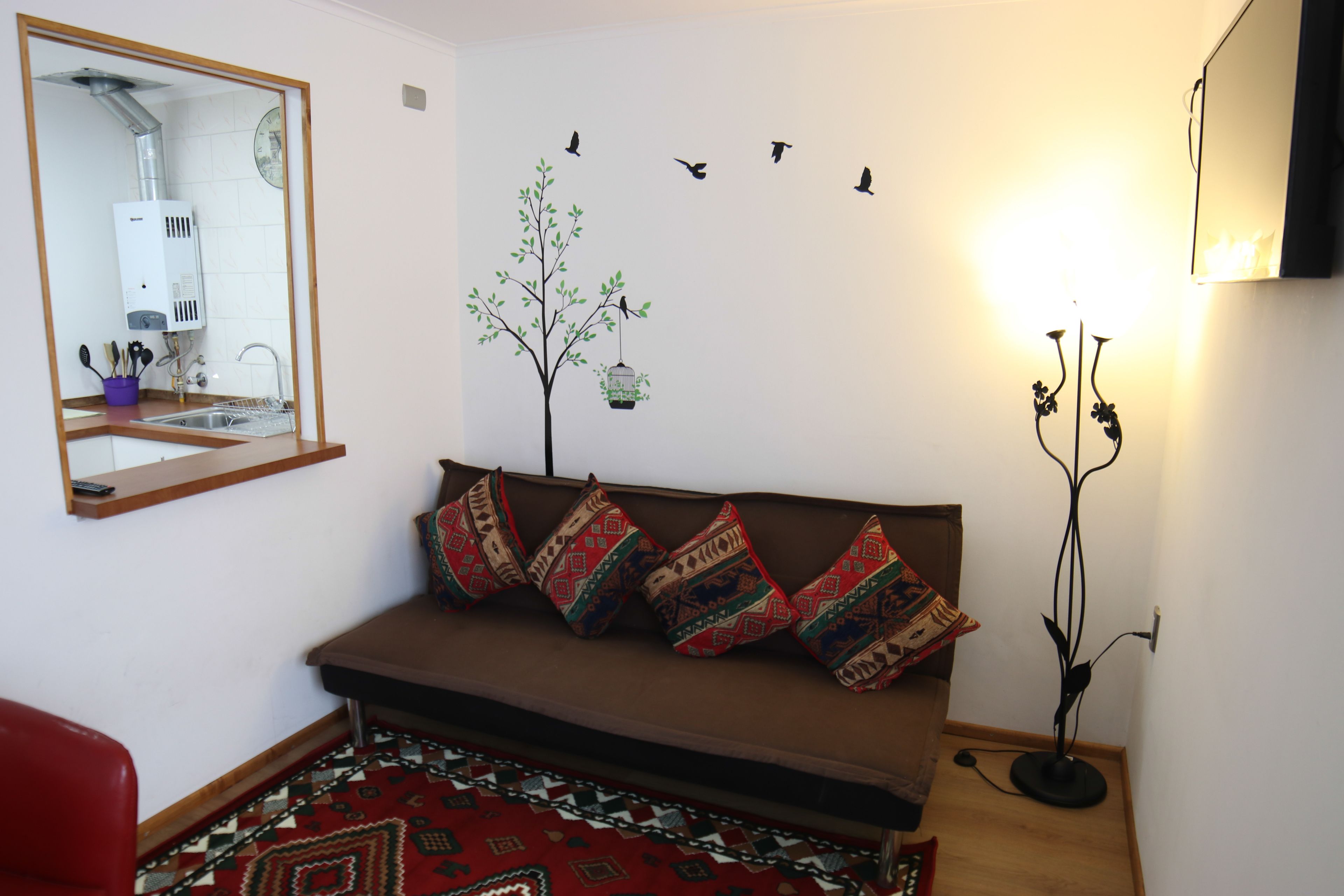 A room with a brown sofa, patterned cushions, a wall-mounted mirror, a radiator, a wall clock, a wall-mounted light, and a decorative plant.