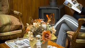Een gezellige kamer met een open haard, een houten stoel, een tafel met kranten en een bloemenvaas.