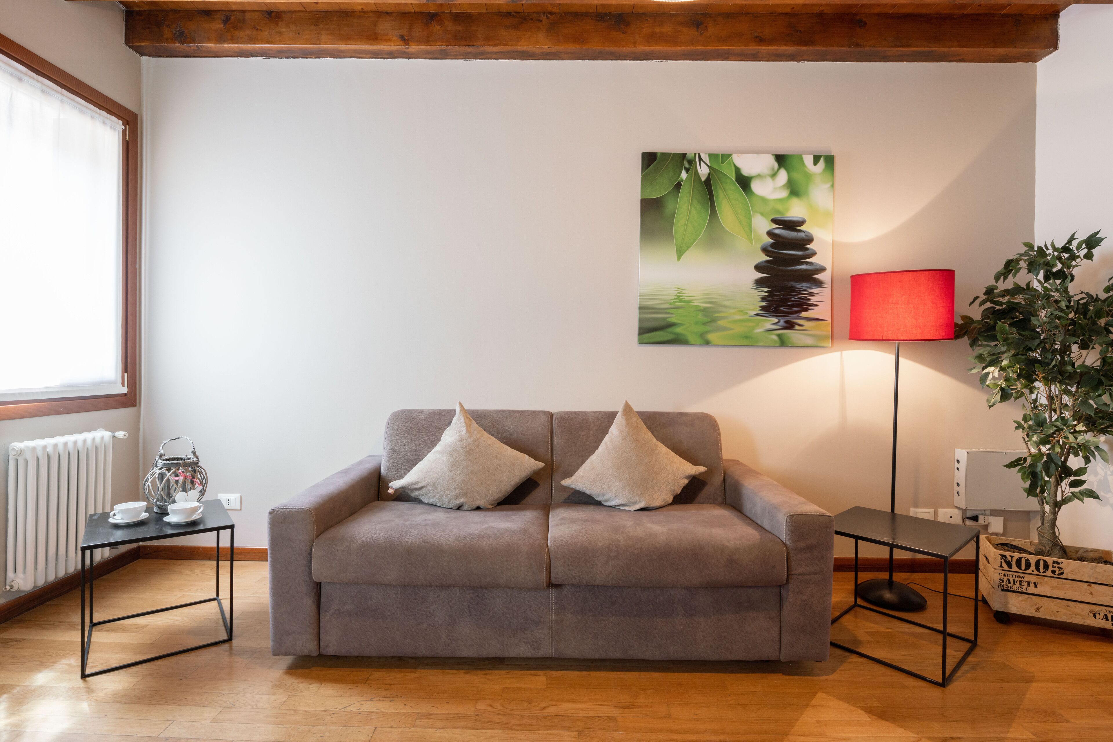 Una sala de estar con un sofá gris, dos almohadas blancas, una lámpara de pie roja, un cuadro de un árbol y una planta en maceta.