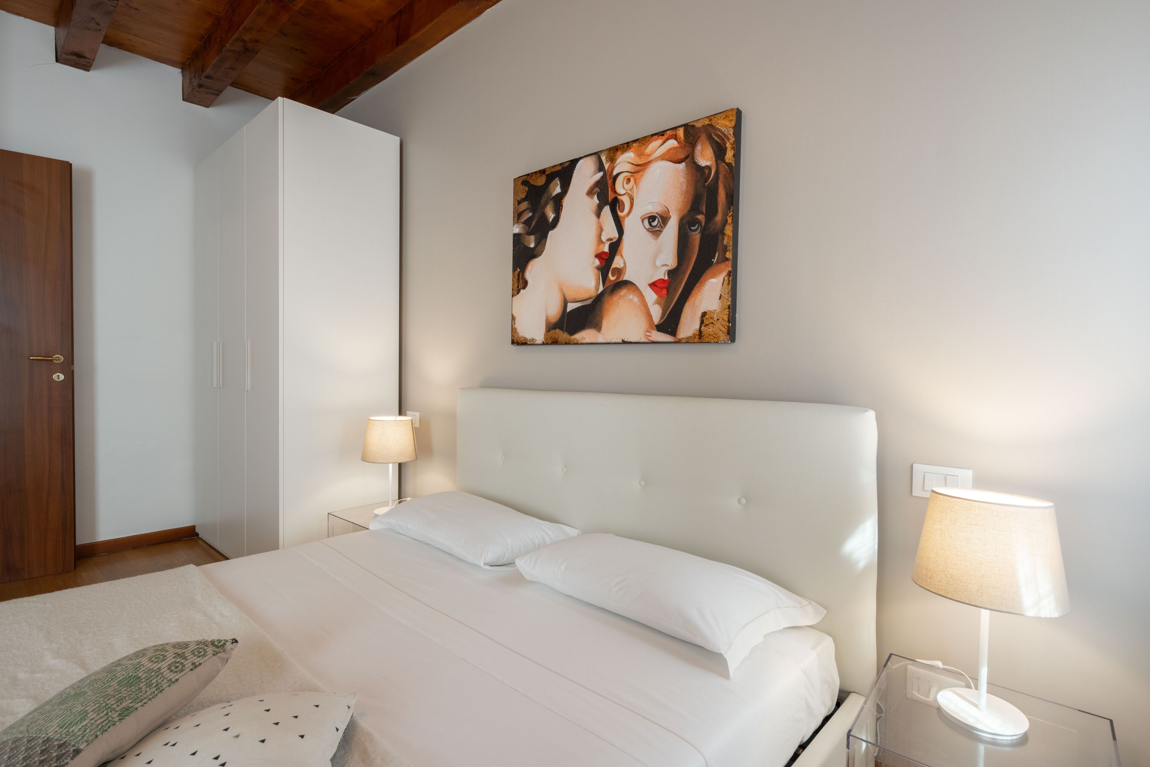 Ein Schlafzimmer mit einem Bett, zwei Nachttischlampen, einem Bild an der Wand und einer Holztür.