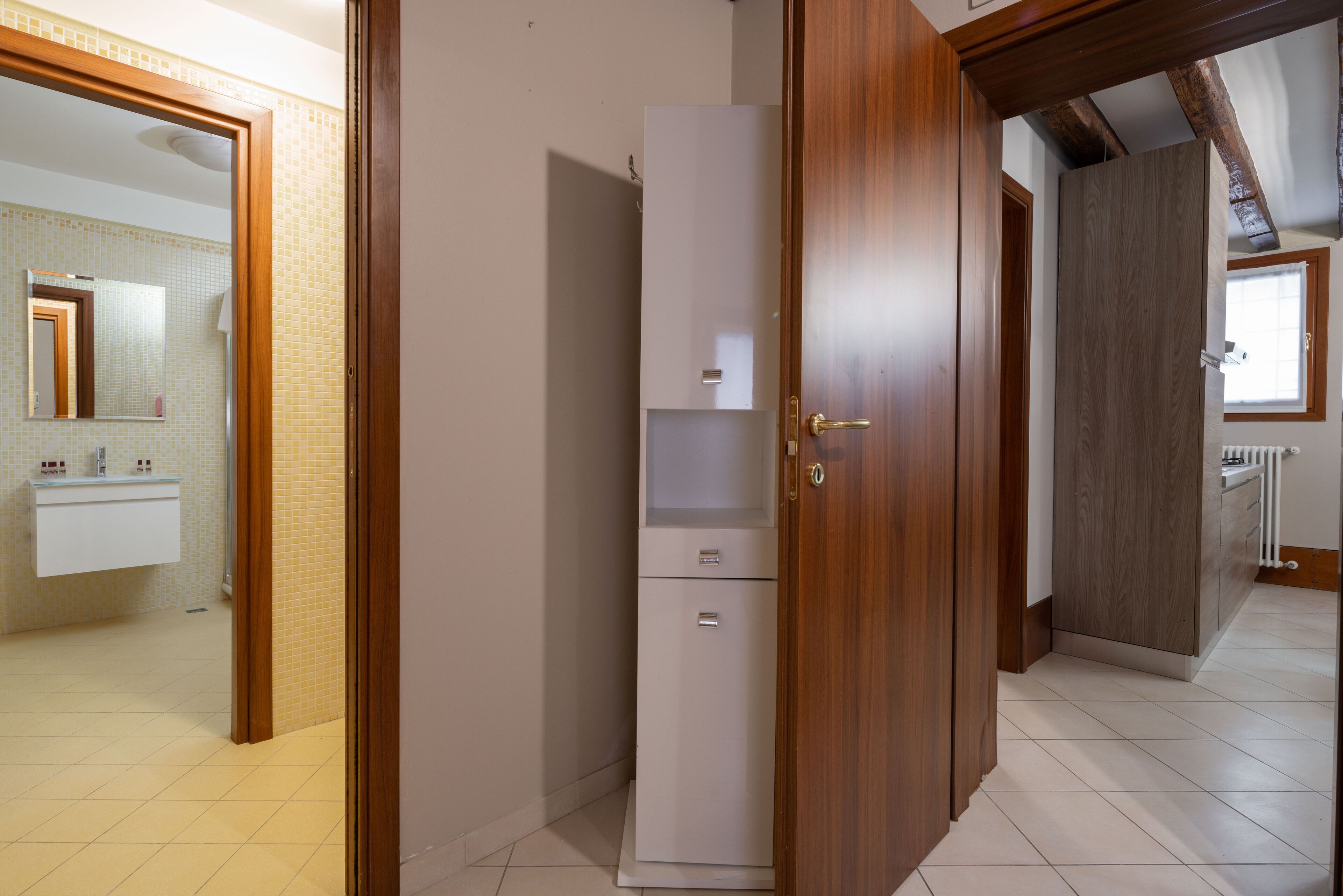 Un couloir avec un réfrigérateur blanc, des portes en bois et une salle de bain visible au fond.
