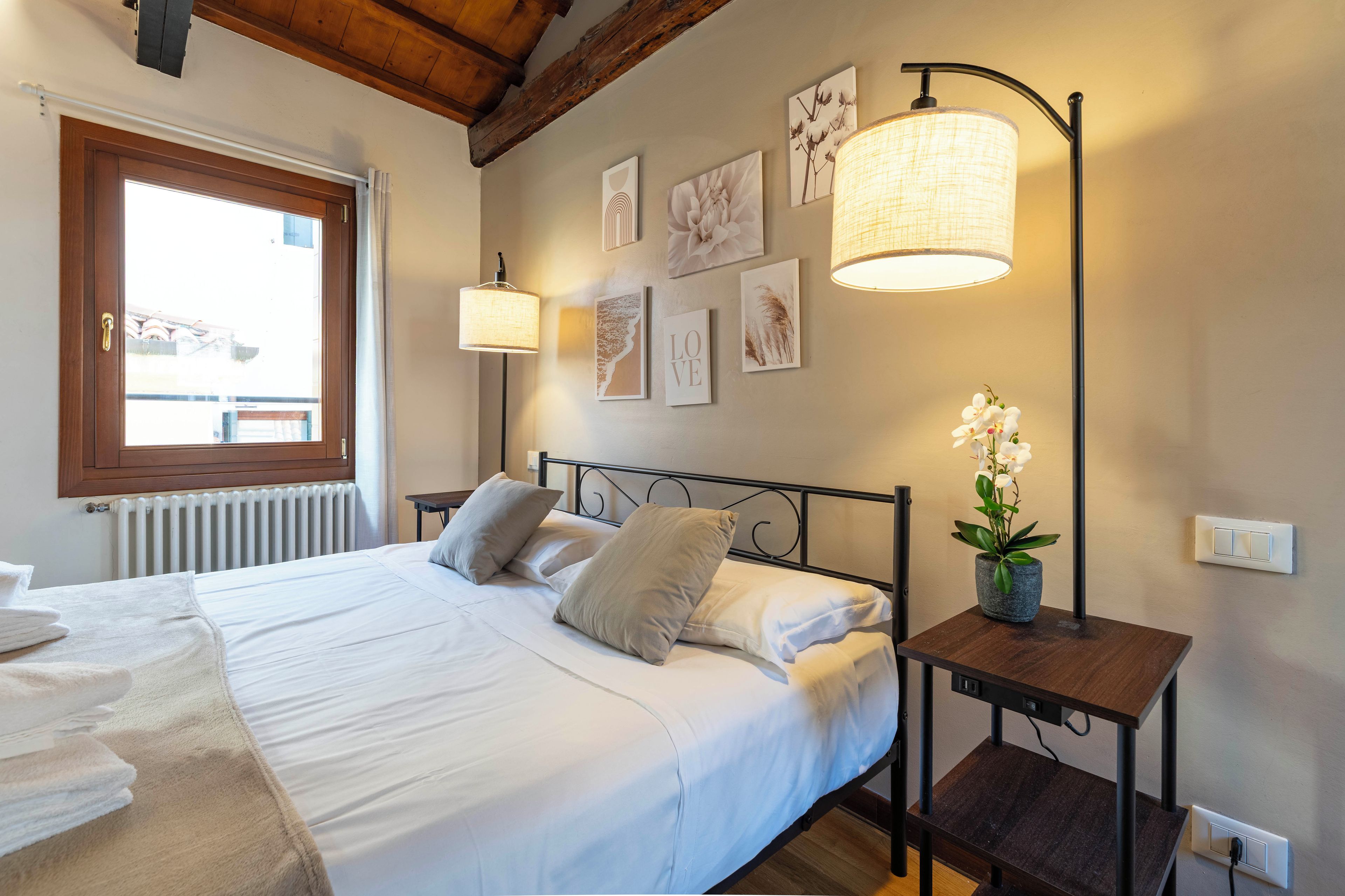 Een net opgemaakt bed met kussens, een nachtkastje met een lamp en een muur met ingelijste foto's.