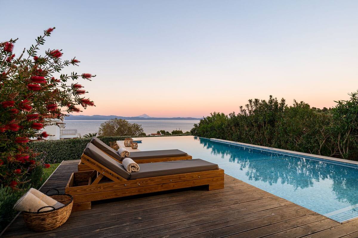 An infinity pool with wooden loungers and a view of the sea at sunset.