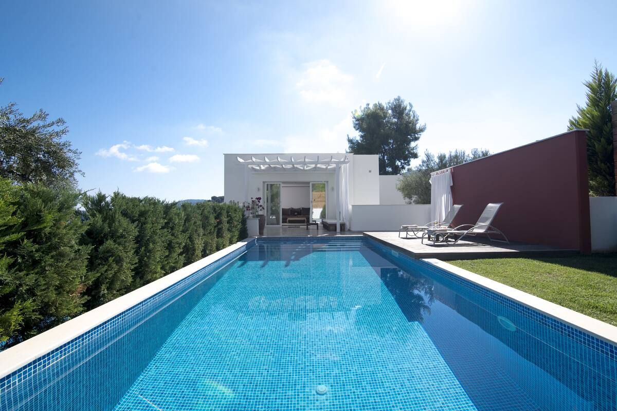 A rectangular swimming pool with a clear blue tile surface, surrounded by a white pool deck and a red building.