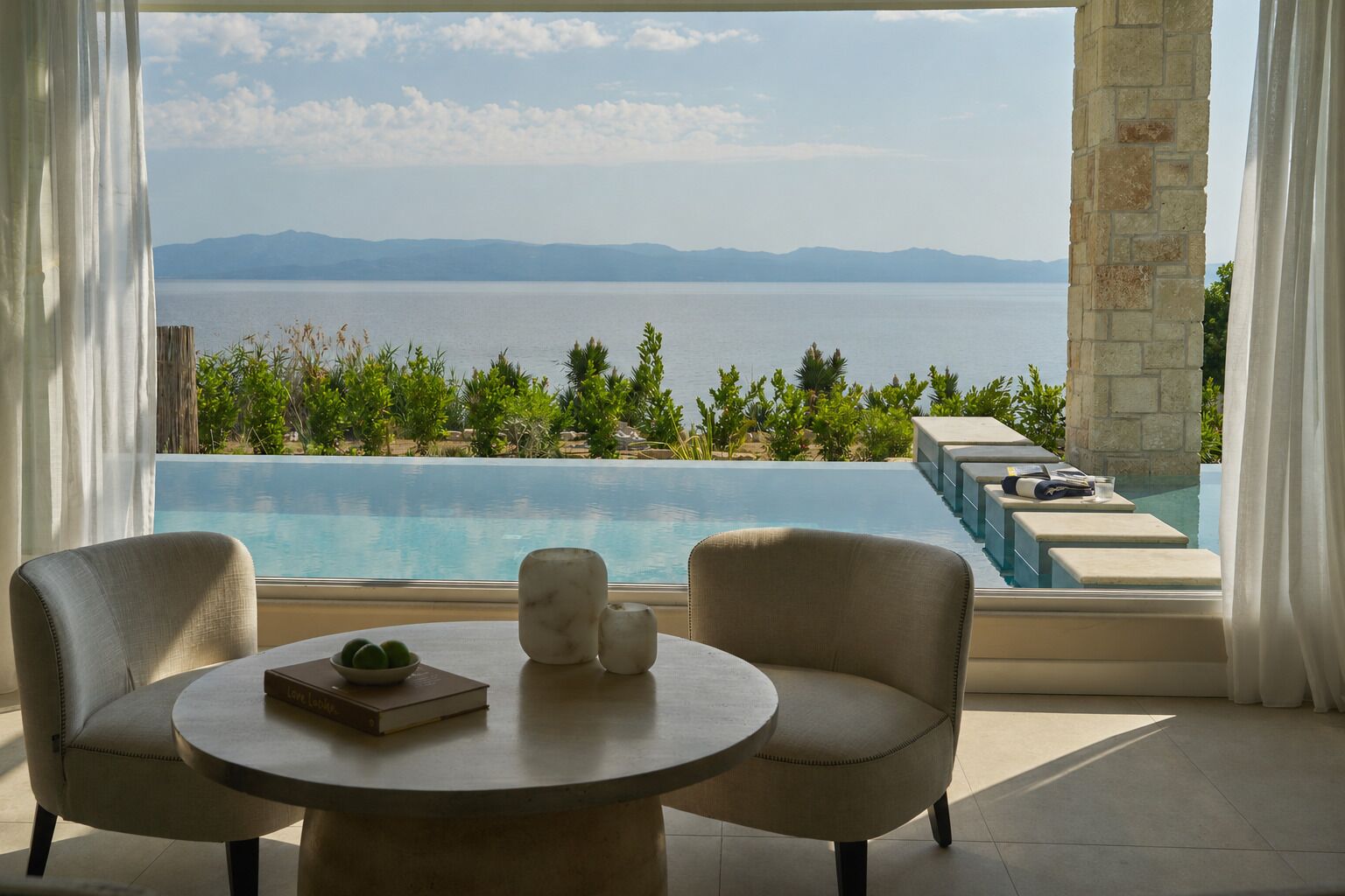 A room with a view of a large body of water, a pool, and a table with chairs.