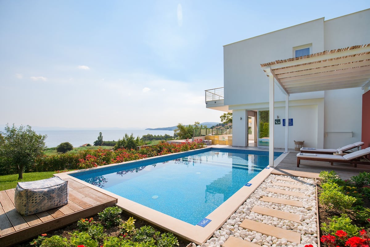 A modern outdoor pool area with a clear view of the sea, surrounded by a wooden deck and a garden with red flowers.