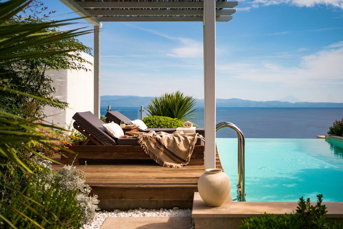 A wooden deck with lounge chairs and a towel, overlooking a clear blue pool with a view of the sea and mountains.
