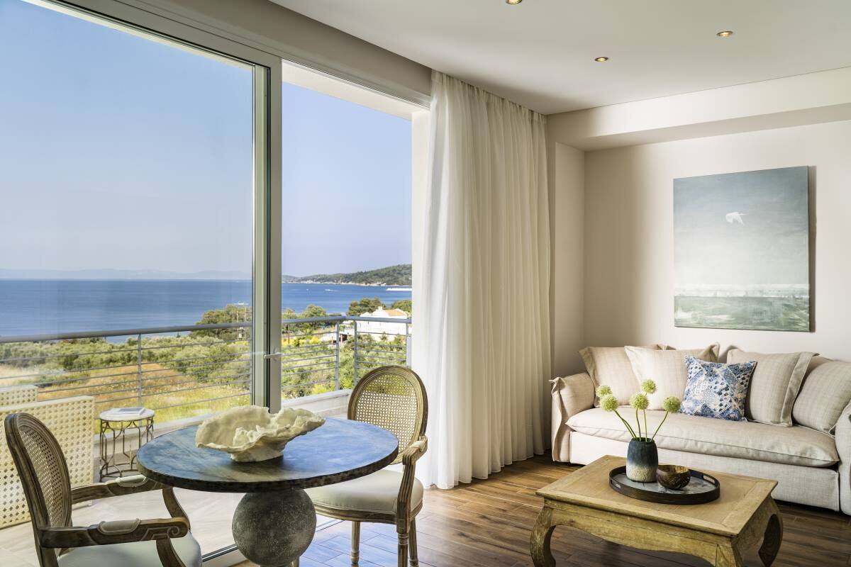 A spacious living room with a large window offering a view of the sea and greenery. There is a round table with chairs, a sofa, and a coffee table with decorative items.