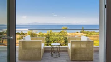 A balcony with wicker furniture and a small round table, overlooking a coastal landscape with mountains in the distance.