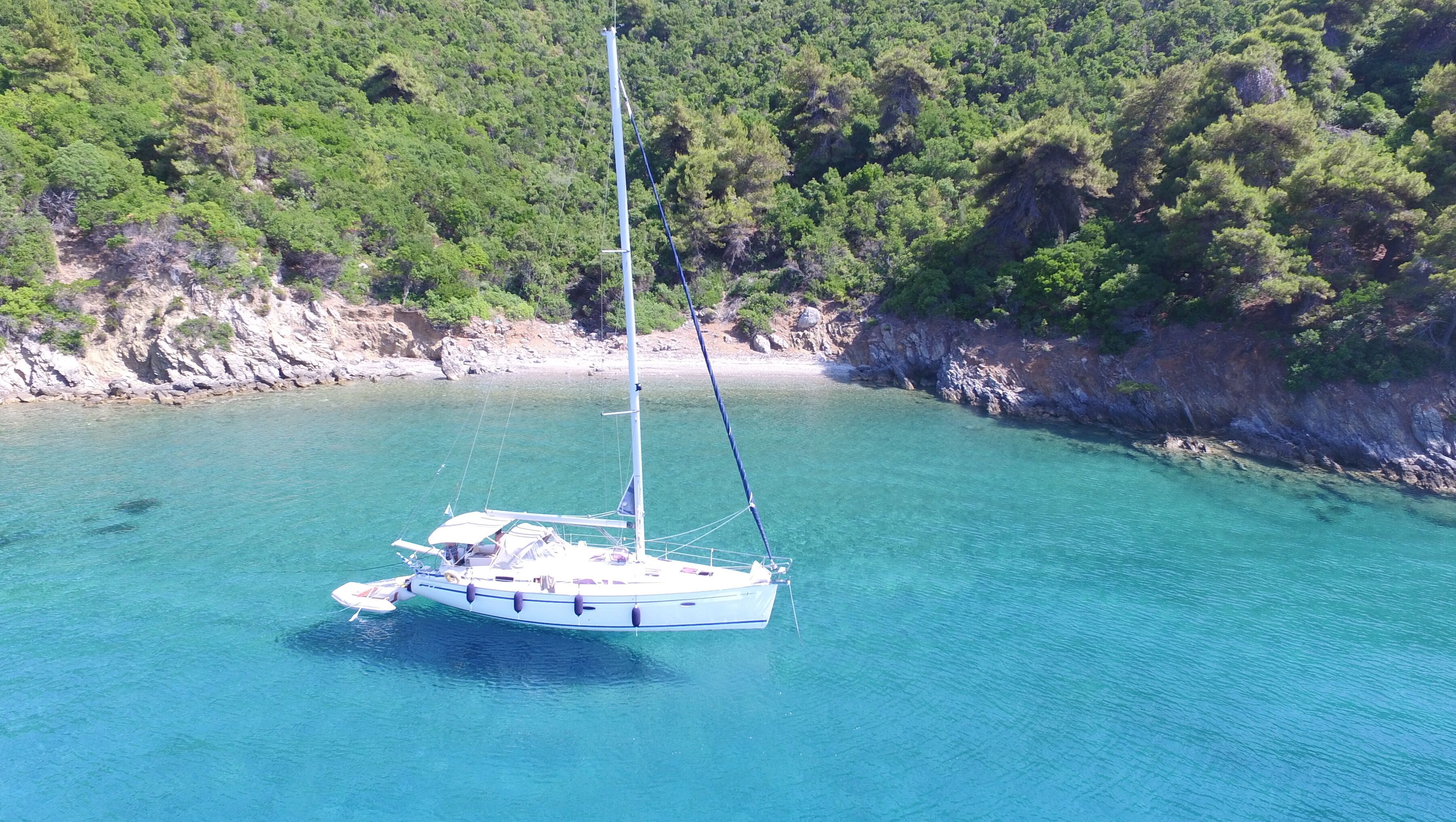 A sailboat anchored in clear turquoise waters with a rocky coastline and dense green forest in the background.