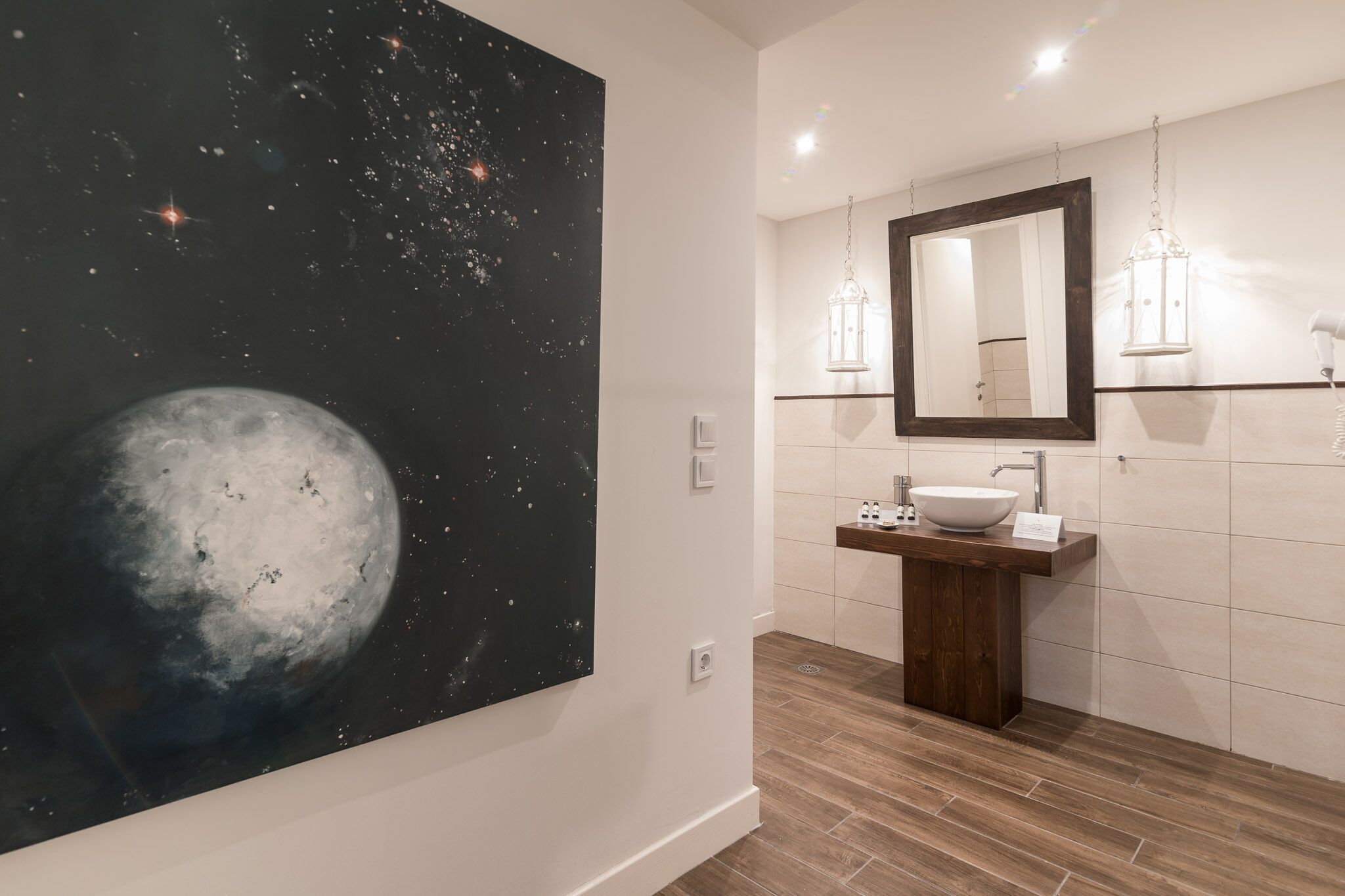 Een badkamer met een grote muurschildering van een maan, een houten wastafelmeubel met een witte wasbak en twee wandlampen.